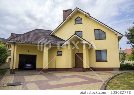 New nice yellow two-story residential cottage with shingle roof, plastic windows, high chimney, paved yard, car in attached garage on blue sky background. Real estate property and prosperity concept. New nice yellow two-story residential cottage with shingle roof, plastic windows, high chimney, paved yard, car in attached garage on blue sky background. Real estate property and prosperity concept. 138082778