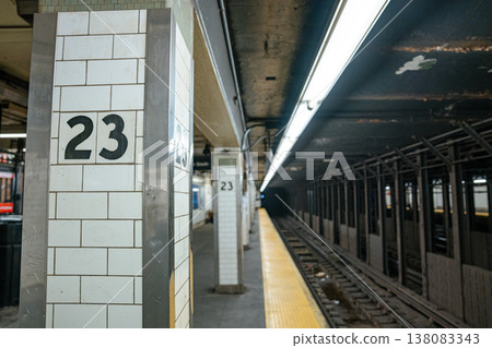 A white tiled column marked 23 stands beside underground subway tracks in Manhattan, New York City. Fluorescent lights and steel framework define the NYC metro transportation system. 138083343
