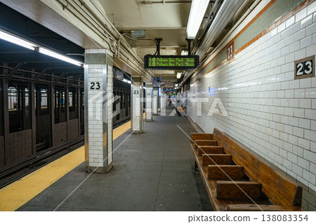 The 23 Street subway station platform in Manhattan features white tiled walls and wooden benches. Digital signs display train arrival times in the New York City metro system. 138083354