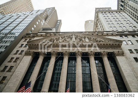 The New York Stock Exchange building rises beneath tall skyscrapers in Manhattan financial district. Classical columns and sculptures highlight the historic Wall Street facade. 138083362