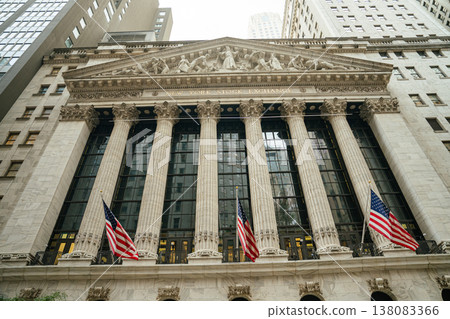 Front view of the New York Stock Exchange building with tall classical columns on Wall Street. American flags wave above the entrance representing financial power in Manhattan. 138083366