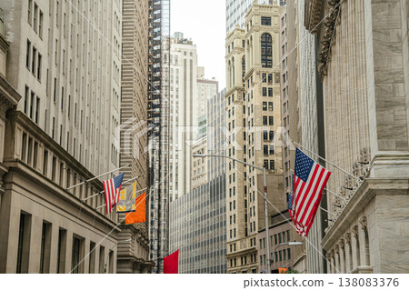 Historic financial buildings and skyscrapers line Wall Street in New York City financial district. Multiple American flags hang along the street representing business, finance, and the United States Historic financial buildings and skyscrapers line Wall Street in New York City financial district. Multiple American flags hang along the street representing business, finance, and the United States 138083376