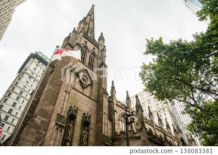 A tall Gothic church spire rises between modern and historic skyscrapers in New York City. The ornate stone architecture contrasts with surrounding office towers in downtown Manhattan. 138083381