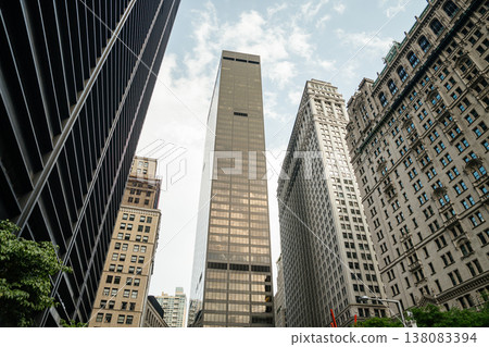 A dramatic low angle view shows tall office towers and skyscrapers in Manhattan New York. Glass and stone buildings reflect light in the busy downtown financial district. 138083394