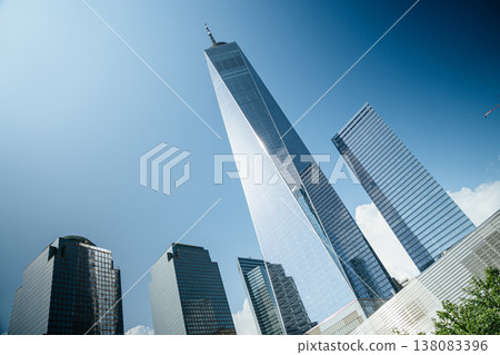 Angled perspective of One World Trade Center towering over surrounding skyscrapers in Manhattan. The modern glass facade reflects sunlight against a bright blue sky. 138083396