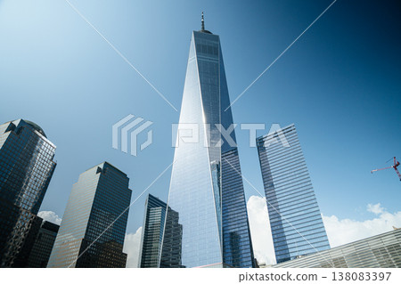 One World Trade Center dominates the Lower Manhattan skyline with reflective glass architecture. The tall tower stands among modern office buildings under a clear blue sky in New York City. One World Trade Center dominates the Lower Manhattan skyline with reflective glass architecture. The tall tower stands among modern office buildings under a clear blue sky in New York City. 138083397