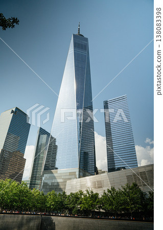 One World Trade Center rises above the 9 11 Memorial plaza in Lower Manhattan, New York City. The modern glass skyscraper stands tall against a blue sky surrounded by trees and downtown buildings. One World Trade Center rises above the 9 11 Memorial plaza in Lower Manhattan, New York City. The modern glass skyscraper stands tall against a blue sky surrounded by trees and downtown buildings. 138083398