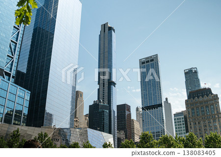 Modern glass skyscrapers rise above green trees in Lower Manhattan under a clear blue sky. The financial district skyline blends contemporary towers with historic buildings in New York City. Modern glass skyscrapers rise above green trees in Lower Manhattan under a clear blue sky. The financial district skyline blends contemporary towers with historic buildings in New York City. 138083405