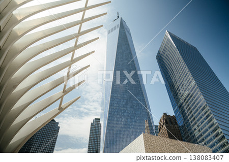 One World Trade Center towers over Lower Manhattan beside the white architectural ribs of the Oculus. The modern glass skyscraper reflects the blue sky in New York City's financial district. One World Trade Center towers over Lower Manhattan beside the white architectural ribs of the Oculus. The modern glass skyscraper reflects the blue sky in New York City's financial district. 138083407