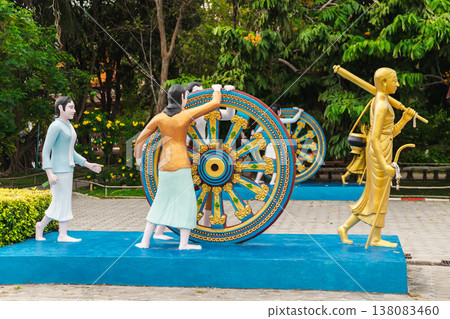 Colorful sculpture of people pushing Dharma wheel behind monk statue in temple garden in Thailand Colorful sculpture of people pushing Dharma wheel behind monk statue in temple garden in Thailand 138083460