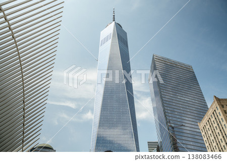 One World Trade Center towers into the blue sky in Lower Manhattan, New York City. The modern glass skyscraper reflects clouds above the downtown financial district. 138083466