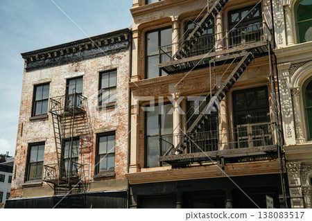 Brick and cast iron buildings with fire escapes line a street in SoHo, New York City. Detailed facades and large windows reflect the historic character of Manhattan architecture. 138083517