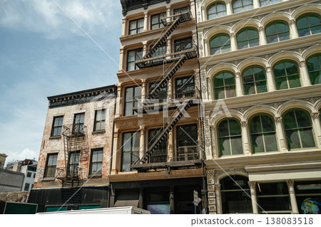 Cast iron buildings with black metal fire escapes stand along a SoHo street in New York City. Ornate facades and large windows showcase classic Manhattan architectural style. 138083518