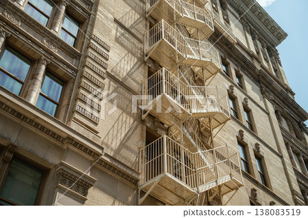A historic New York City building features a white metal fire escape against a detailed beige brick facade. Strong sunlight creates dramatic shadows highlighting classic Manhattan architecture. 138083519