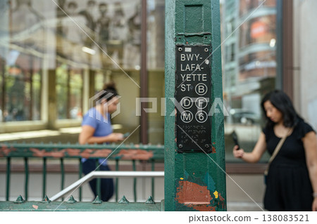 A Broadway Lafayette Street subway sign displays multiple train lines at a New York City entrance. Pedestrians using smartphones pass by showing modern city commuting behavior. A Broadway Lafayette Street subway sign displays multiple train lines at a New York City entrance. Pedestrians using smartphones pass by showing modern city commuting behavior. 138083521