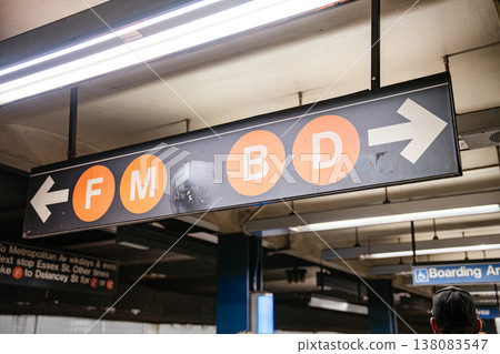 Overhead subway direction sign showing F M B and D train lines at Broadway Lafayette Street station in New York City. Underground metro scene with fluorescent lighting and urban transit infrastructure Overhead subway direction sign showing F M B and D train lines at Broadway Lafayette Street station in New York City. Underground metro scene with fluorescent lighting and urban transit infrastructure 138083547