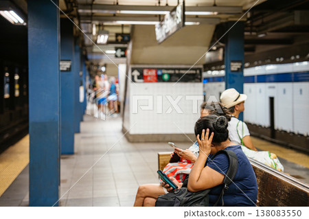Woman using smartphone while sitting on bench at Broadway Lafayette Street subway station in New York City. Urban underground metro platform scene showing daily commute, tiled walls, and public Woman using smartphone while sitting on bench at Broadway Lafayette Street subway station in New York City. Urban underground metro platform scene showing daily commute, tiled walls, and public 138083550