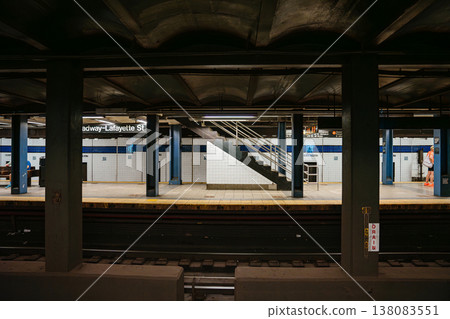 Symmetrical underground view of Broadway Lafayette Street subway station platform in New York City. Urban metro scene with staircase, tiled walls, railway tracks, and industrial steel columns. Symmetrical underground view of Broadway Lafayette Street subway station platform in New York City. Urban metro scene with staircase, tiled walls, railway tracks, and industrial steel columns. 138083551