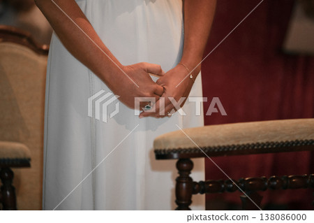 Bride standing with clasped hands near vintage chairs before ceremony in a church Bride standing with clasped hands near vintage chairs before ceremony in a church 138086000