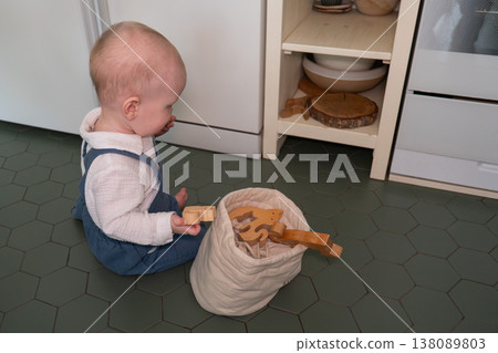 Baby plays with wooden toys next to fabric storage bag on tiled floor in home interior, everyday play and early development moment. Child development, parenting content, family life, lifestyle 138089803
