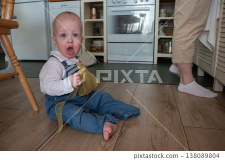 Baby sits on kitchen floor holding silicone bib and looks surprised at camera, everyday feeding routine in home interior. Child development, parenting content, family life, lifestyle marketing, home 138089804