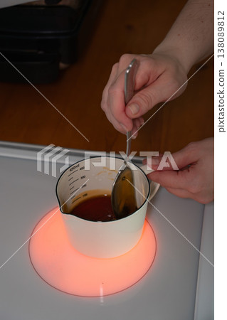 Hands stir honey in measuring cup on heated stovetop during baking preparation in home kitchen. Cooking process, ingredient preparation, homemade pastry, lifestyle content, kitchen 138089812
