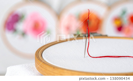 A thin silver needle is moving through bright crimson silk thread and white fabric, showing a detail of the embroidery process. Background features colorful floral designs. 138090434
