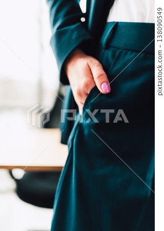 Person stands with hand in pocket wearing dark suit in office setting during daytime with desk and chair visible Person stands with hand in pocket wearing dark suit in office setting during daytime with desk and chair visible 138090549