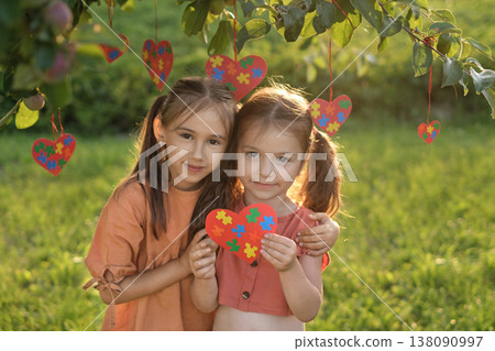 Children are standing in the park under a tree decorated with hearts from puzzle pieces, showing a postcard for autism Day with love and care Children are standing in the park under a tree decorated with hearts from puzzle pieces, showing a postcard for autism Day with love and care 138090997