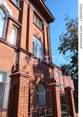 Facade of an old red brick building with arched windows and decorative white accents, partially shaded by trees and sunlight reflections. The image is suitable for topics about building restoration Facade of an old red brick building with arched windows and decorative white accents, partially shaded by trees and sunlight reflections. The image is suitable for topics about building restoration 138091603