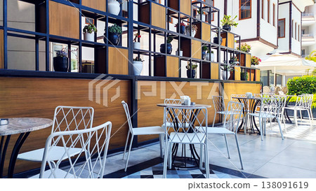 Minimalist caf terrace with empty tables and chairs in a bright hotel courtyard surrounded by plants. The peaceful travel scene reflects vacation relaxation, outdoor dining and sunny resort mornings. Minimalist caf terrace with empty tables and chairs in a bright hotel courtyard surrounded by plants. The peaceful travel scene reflects vacation relaxation, outdoor dining and sunny resort mornings. 138091619