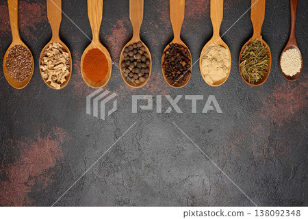 Various dried spices, seeds and roots in wooden spoons arranged in a row on a dark textured surface Various dried spices, seeds and roots in wooden spoons arranged in a row on a dark textured surface 138092348