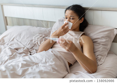 Young black woman lying in bed checking thermometer while holding tissue Young black woman lying in bed checking thermometer while holding tissue 138092666