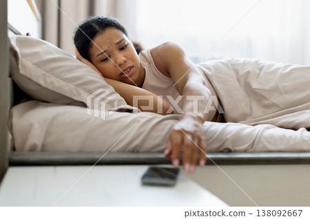 Young black woman lying in bed reaching for smartphone with worried expression Young black woman lying in bed reaching for smartphone with worried expression 138092667