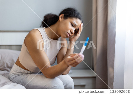 Young black woman sitting on bed holding pregnancy test with worried expression Young black woman sitting on bed holding pregnancy test with worried expression 138092668