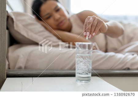 Young black woman lying in bed taking medicine with glass of water 138092670