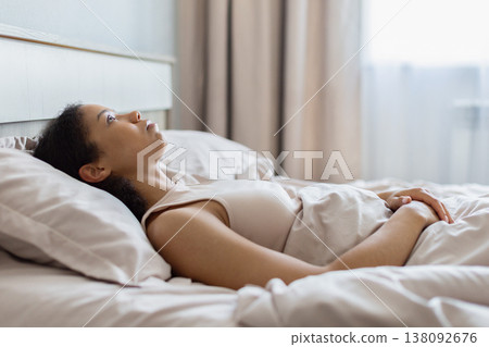 Young black woman lying on bed staring at ceiling with thoughtful expression Young black woman lying on bed staring at ceiling with thoughtful expression 138092676