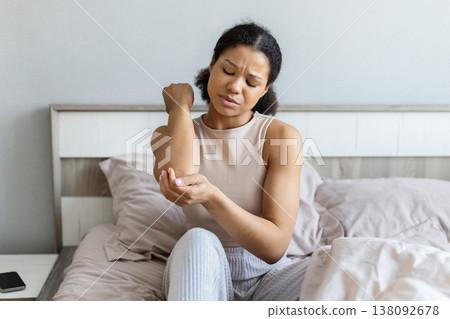Young black woman sitting on bed holding elbow in pain with discomfort 138092678