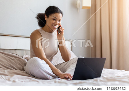 Young black woman sitting on bed talking on smartphone and using laptop 138092682