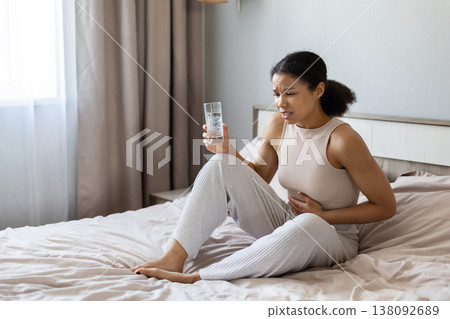 Young black woman sitting on bed holding stomach and glass of water Young black woman sitting on bed holding stomach and glass of water 138092689