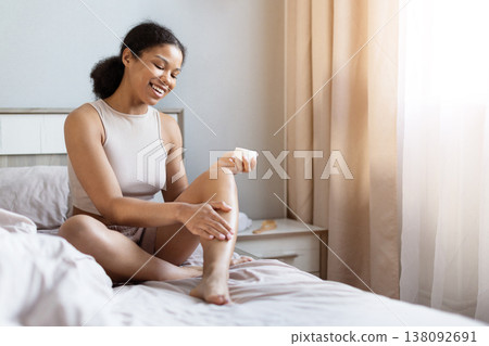 Young black woman sitting on bed applying skincare cream to leg, smiling Young black woman sitting on bed applying skincare cream to leg, smiling 138092691