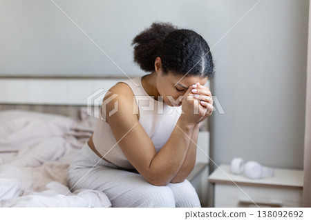 Upset black woman sitting on bed with hands clasped and eyes closed Upset black woman sitting on bed with hands clasped and eyes closed 138092692