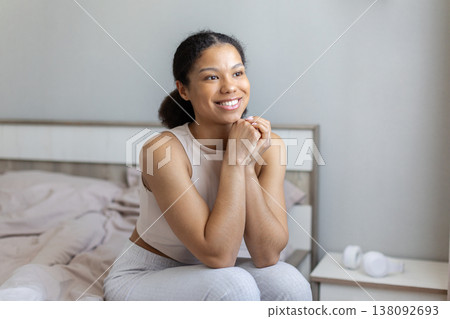 Young black woman sitting on bed smiling and looking ahead Young black woman sitting on bed smiling and looking ahead 138092693