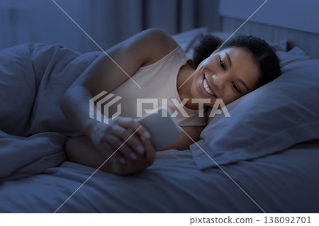 Young black woman lying in bed at night smiling while using smartphone Young black woman lying in bed at night smiling while using smartphone 138092701