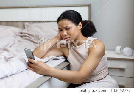 Young black woman sitting beside bed looking at smartphone with worried expression Young black woman sitting beside bed looking at smartphone with worried expression 138092722