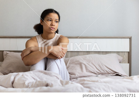 Black woman sitting on bed hugging knees, looking away with sad expression Black woman sitting on bed hugging knees, looking away with sad expression 138092731