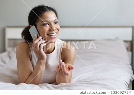 Black woman talking on smartphone while lying on bed, smiling and gesturing Black woman talking on smartphone while lying on bed, smiling and gesturing 138092734