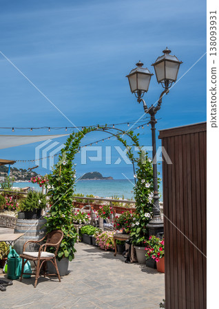 Alassio seaside terrace with a green floral arch, antique street lamp and potted plants framing a sunny view of gallinara island and the blue mediterranean horizon 138093931