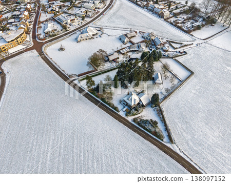 Top down aerial view of a snowy suburban neighborhood and frost covered fields. A clean winter landscape featuring modern houses, garden plots, and a long road slicing through the white terrain. 138097152