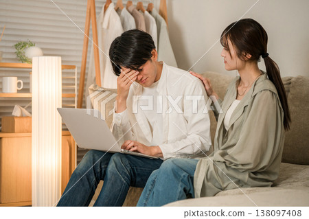A wife looks anxiously at her husband working on a computer on the sofa in the living room (overtime, taking work home, hidden overtime) A wife looks anxiously at her husband working on a computer on the sofa in the living room (overtime, taking work home, hidden overtime) 138097408